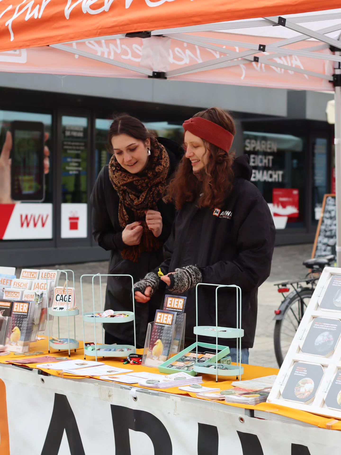 Zwei ARIWA-Aktivistinnen hinter dem Infostand-Tisch unter dem orangefarbenen Pavillon. Auf dem Tisch Broschüren zu Themen wie Eier, Milch, Fische, Fleisch, Leder und Jagd sowie Buttons in Ständern. Beide schauen auf etwas auf dem Tisch. Im Hintergrund ein WVV-Gebäude.