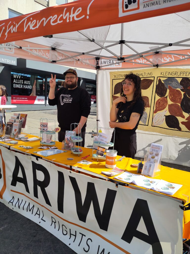 Zwei lachende ARIWA-Aktivist:innen hinter dem Infostand mit gelbem Tuch und ARIWA-Banner. Links ein Mann mit Basecap zeigt Victory-Zeichen. Im Hintergrund ein handgemaltes Schaubild zu Haltungsformen. Auf dem Tisch Broschüren, Buttons und eine Spendenbox.