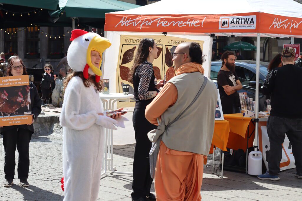 ARIWA-Infostand auf einem Würzburger Stadtplatz bei Sonnenschein. Im Vordergrund eine Aktivistin im weißen Hühnerkostüm im Gespräch mit einer Aktivist:in, daneben ein Mann in traditioneller orangefarbener Robe. Links hält eine weitere Aktivistin das Legehennen-Schild. Im Hintergrund der ARIWA-Pavillon mit Haltungsformen-Schaubild.