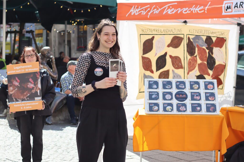 Lächelnde ARIWA-Aktivistin hält Flyer vor dem Stand mit Eier-Infodisplay und Haltungsformen-Schaubild. Links im Hintergrund Aktivistin mit Legehennen-Schild. Sonniges Wetter auf einem belebten Stadtplatz.