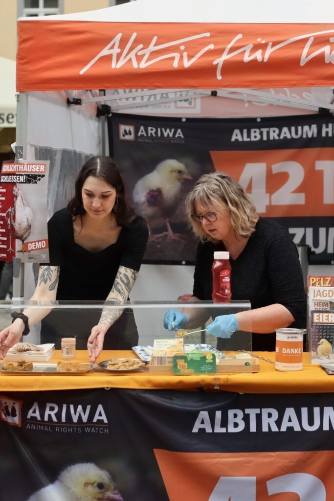 Zwei Aktivistinnen richten am ARIWA-Infostand vegane Kostproben her: eine bereitet Häppchen mit Zahnstocher vor (Einweghandschuhe), die andere arrangiert Teller. Im Hintergrund das Banner „Albtraum Hühnermast – 42 Tage bis zum Tod!" mit Kükenfoto sowie Broschüren zu Pelz, Jagd, Heimtiere und Eier. Spendenbox auf dem Tisch.
