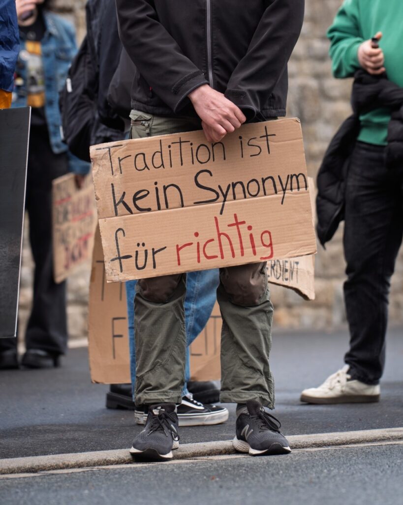 Nahaufnahme eines handgeschriebenen Pappchilds: „Tradition ist kein Synonym für richtig" – „richtig" in Rot hervorgehoben. Im Hintergrund weitere Aktivist:innen mit Schildern.