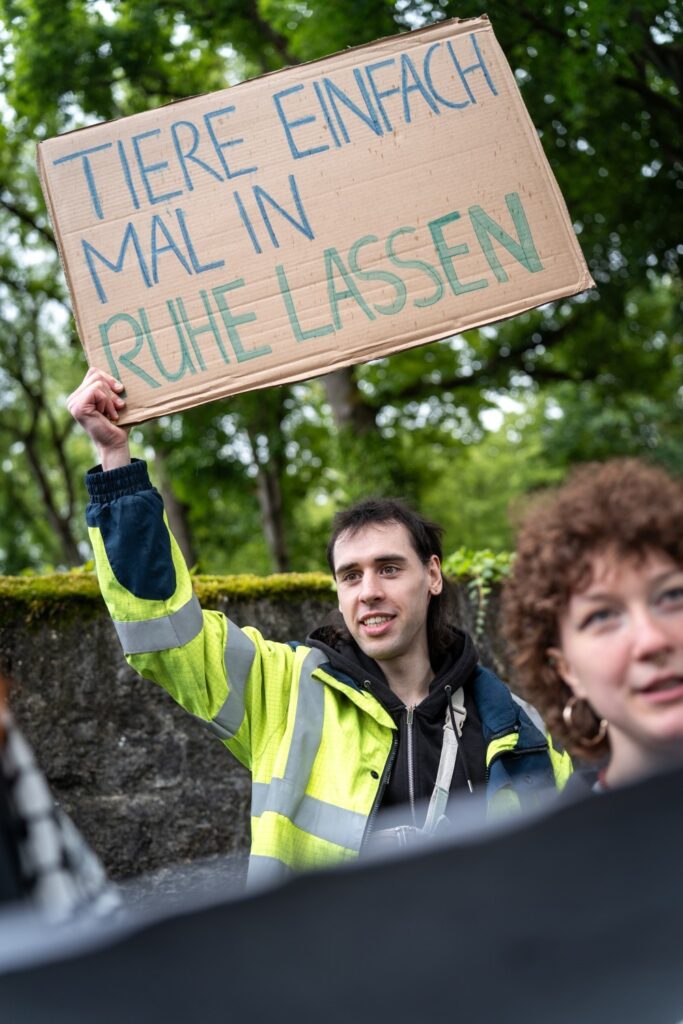 Aktivist in gelber Warnweste streckt ein Pappschild in die Höhe: „Tiere einfach mal in Ruhe lassen". Er lächelt. Neben ihm eine weitere Aktivistin, im Hintergrund Bäume.