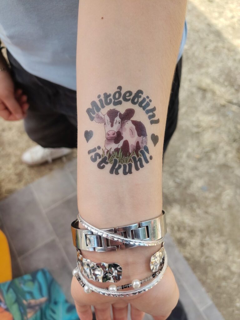 Unterarm einer Person mit Klebetatoo: eine schwarz-weiße Kuh im Kreis mit dem Schriftzug „Mitgefühl ist kuhl!" und Herzchen. Am Handgelenk mehrere Silberarmbänder.