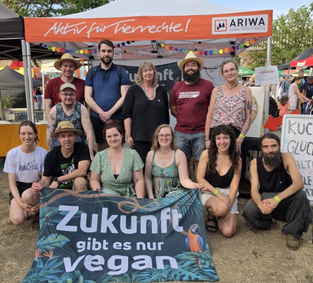 Gruppenfoto von etwa elf Aktivistinnen und Aktivisten der ARIWA Ortsgruppe Würzburg vor ihrem Stand beim Festival Umsonst & Draußen. Im Hintergrund das orangefarbene ARIWA-Pavillon-Zelt mit der Aufschrift „Aktiv für Tierrechte!" sowie ein Schild mit „Kuchen, Glücksrad, Maltisch, Klebe-Tattoos". Die Gruppe hält gemeinsam ein Banner mit der Aufschrift „Zukunft gibt es nur vegan" mit tropischem Papagei-Motiv. Die Personen tragen sommertypische Kleidung, einige mit Strohhüten. Im Hintergrund das bunte Festivalgelände bei Sonnenschein.