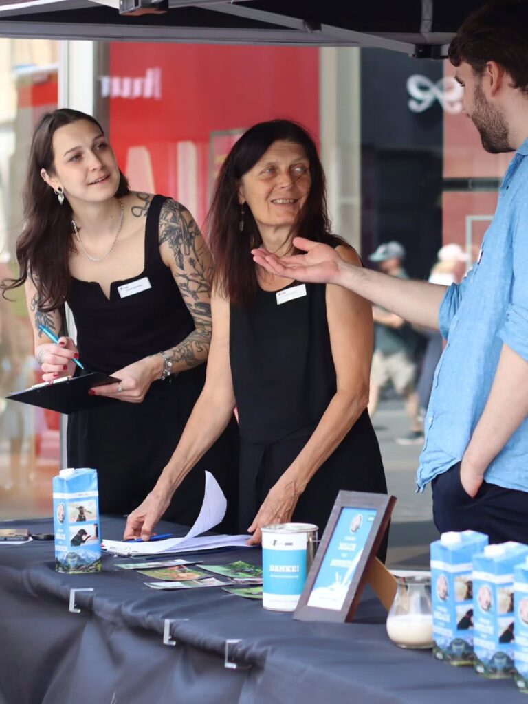Zwei Aktivistinnen in schwarzer Kleidung mit Namensschildern stehen hinter einem Infostand-Tisch und sprechen mit einem Passanten in blauem Hemd, der gestikuliert. Auf dem Tisch liegen Flyer, ein Klemmbrett, eine Spendendose mit „Danke!" und blaue „Milch von Hunden"-Kartons.
