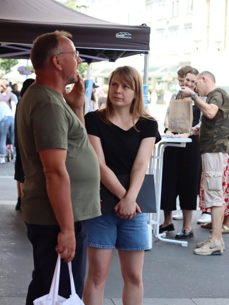 Eine Aktivistin schwarzem Shirt und Jeansshorts, die eine dunkle Mappe hält spricht am Rand eines Infostands mit einem Mann mit Brille im olivgrünen T-Shirt, der skeptisch zuhört. Im Hintergrund weitere Standbesucher unter einem Pavillon in einer Fußgängerzone.