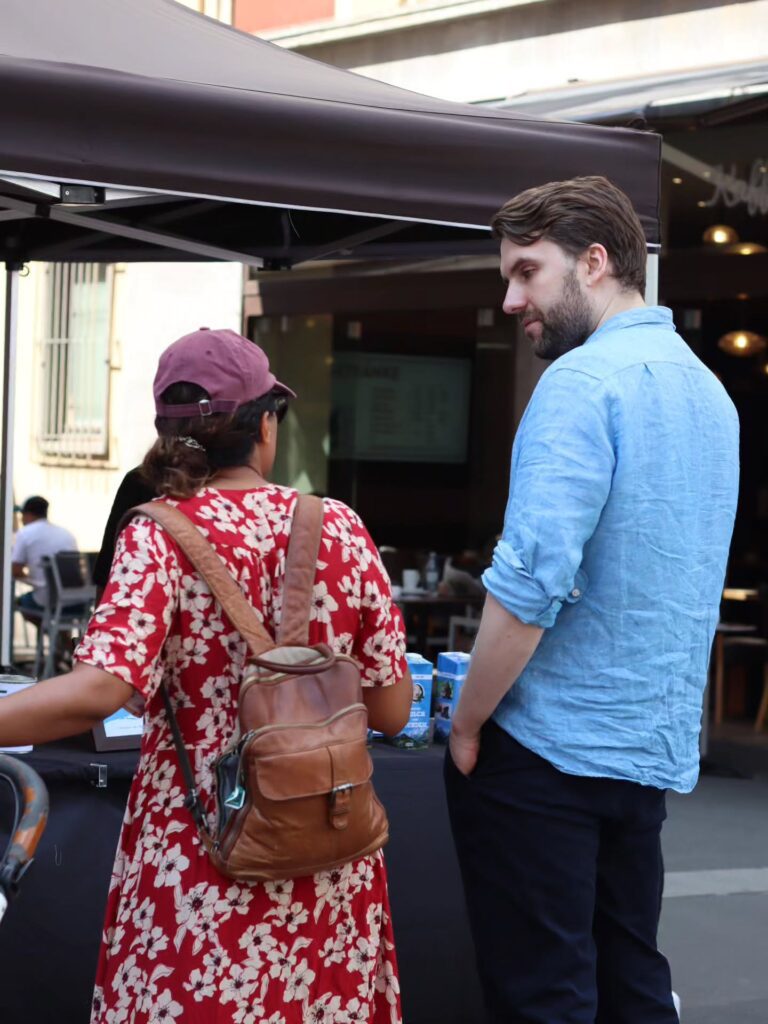 Rückansicht einer Passantin in rotem Blumenkleid mit braunem Lederrucksack, die an einem Infostand mit einem Aktivisten im hellblauen Leinenhemd spricht. Im Hintergrund die blauen „Milch von Hunden"-Kartons und ein Straßencafé.