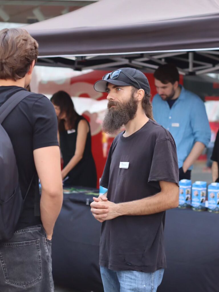 Ein bärtiger Aktivist mit schwarzer Cap und hochgeschobener Sonnenbrille, Namensschild am T-Shirt, steht mit gefalteten Händen unter einem Pavillon und spricht mit einem Standbesucher. Im Hintergrund weitere Aktive und blaue „Milch von Hunden"-Kartons.