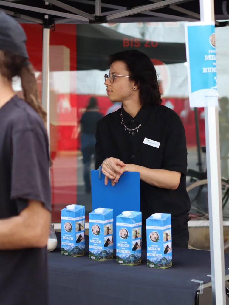 Ein Aktivist in schwarzem Hemd mit Brille, Nasenpiercing und Kette hält ein blaues Klemmbrett. Vor ihm auf dem Tisch vier blaue Kartons „Frische Milch von Hunden" mit Hundekopf-Logo.