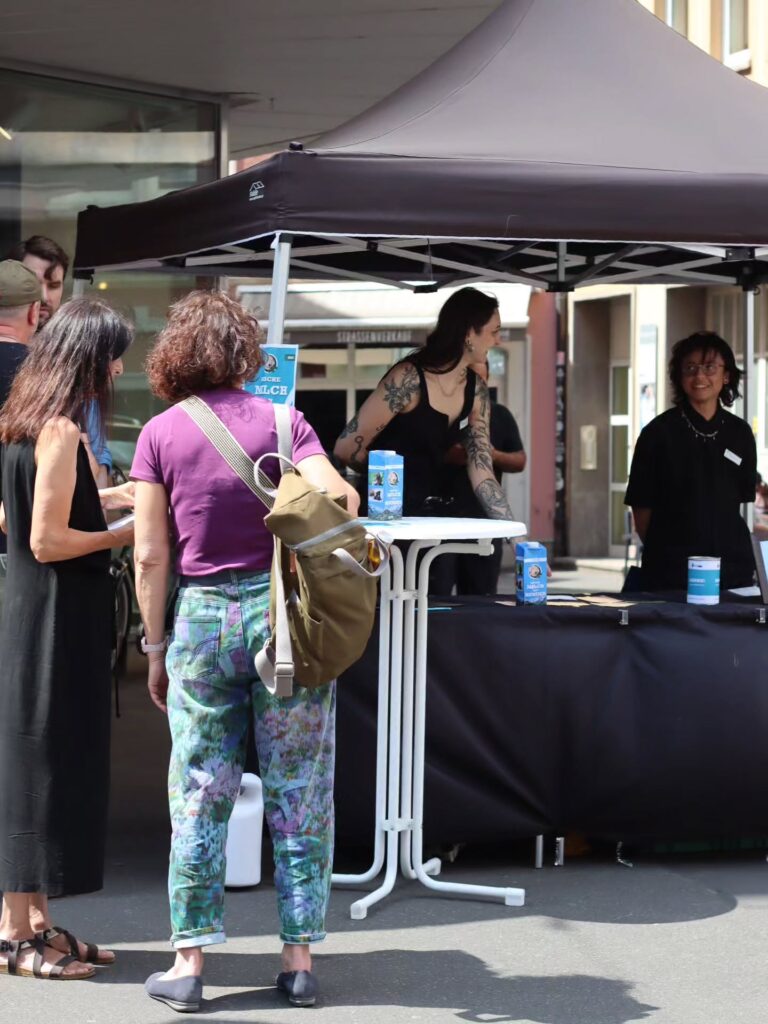 Weitere Ansicht des Infostands in einer Fußgängerzone: Zwei Passantinnen betrachten Material am Tisch, dahinter zwei Aktivistis unter einem schwarzen Pavillon. Auf Tisch und Stehtisch blaue „Milch von Hunden"-Kartons und eine Spendendose.