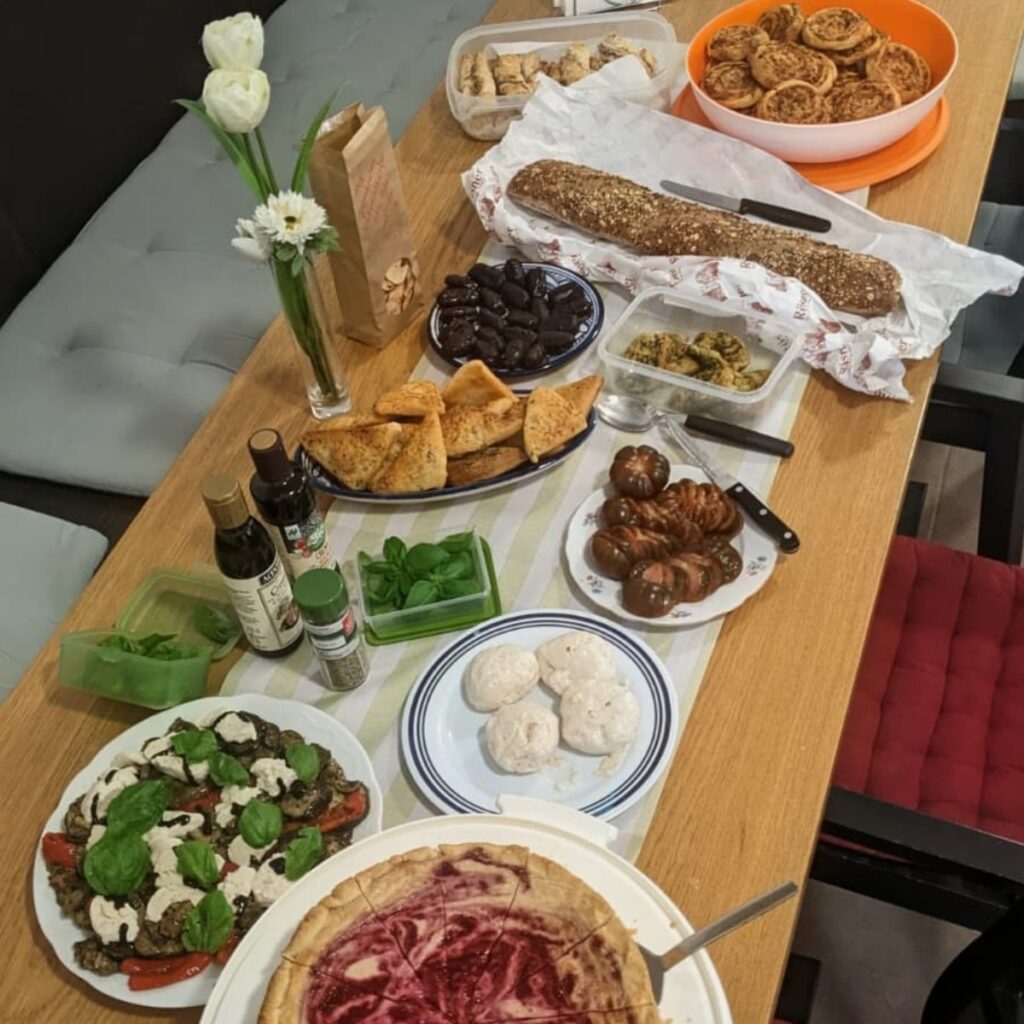 Üppig gedeckter Tisch beim veganen ARIWA-Mitbringbrunch. Zu sehen: Beerenkuchen, veganer Caprese-Salat mit pflanzlichem Mozzarella und Basilikum, Falafel oder Pita-Chips, dunkle Schokoladenoliven, Körner-Baguette, marinierte Zucchini, Schneckengebäck, runde Kokos- oder Marzipankugeln, geschnittene Tomaten sowie Olivenöl und Gewürze. Weiße Blumen als Tischdeko.