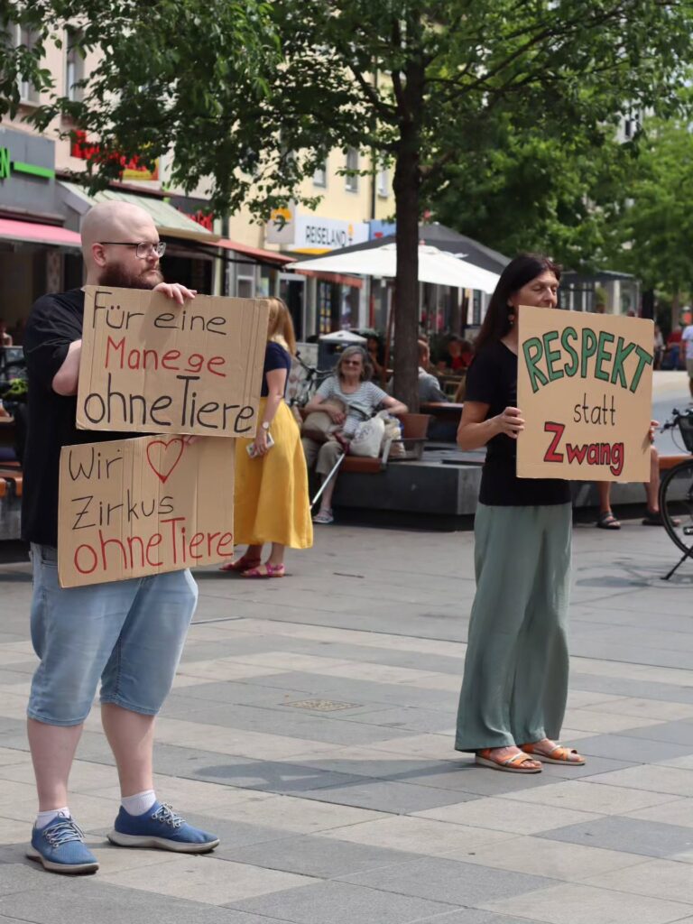 Zwei Aktivist:innen stehen mit Abstand zueinander in einer Fußgängerzone und halten Protestschilder aus Pappe. Links ein Mann mit zwei übereinander gehaltenen Schildern: „Für eine Manege ohne Tiere" und „Wir ♥ Zirkus ohne Tiere". Rechts eine Frau mit dem Schild „Respekt statt Zwang". Im Hintergrund Passant:innen und Straßencafés.