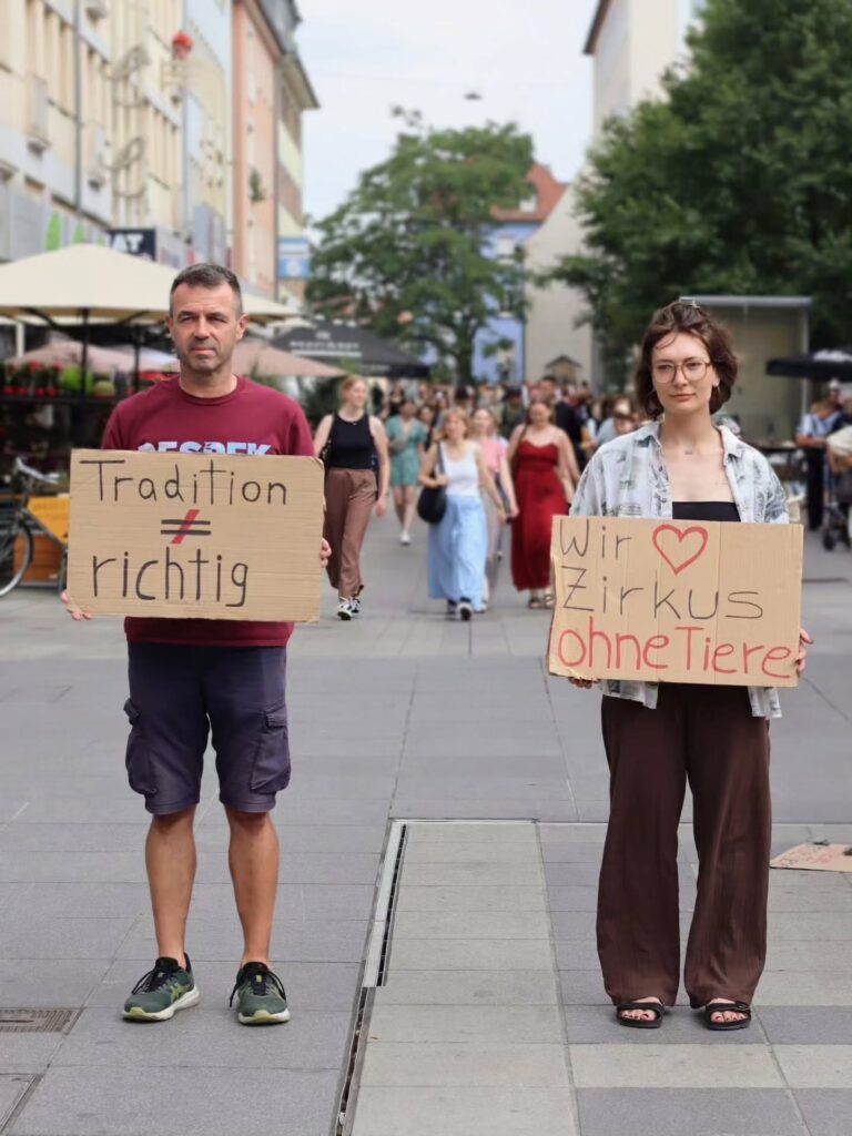 Ein Mann und eine Frau stehen mit Abstand in einer belebten Fußgängerzone und halten Protestschilder. Links: „Tradition ≠ richtig". Rechts: „Wir ♥ Zirkus ohne Tiere". Im Hintergrund viele Passant:innen, Bäume und Straßencafés.