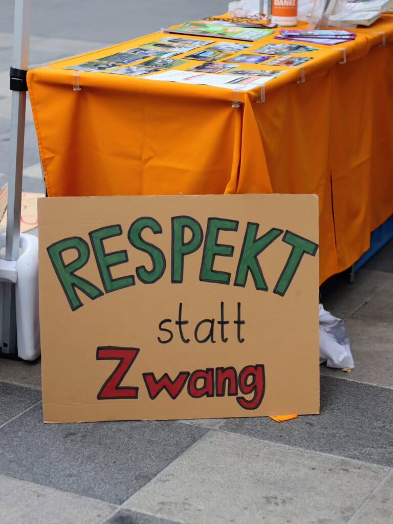 Ein Pappschild mit der Aufschrift „Respekt statt Zwang" in grüner und roter Schrift lehnt vor einem orange gedeckten Infotisch, auf dem Fotos und Flyer ausliegen.
