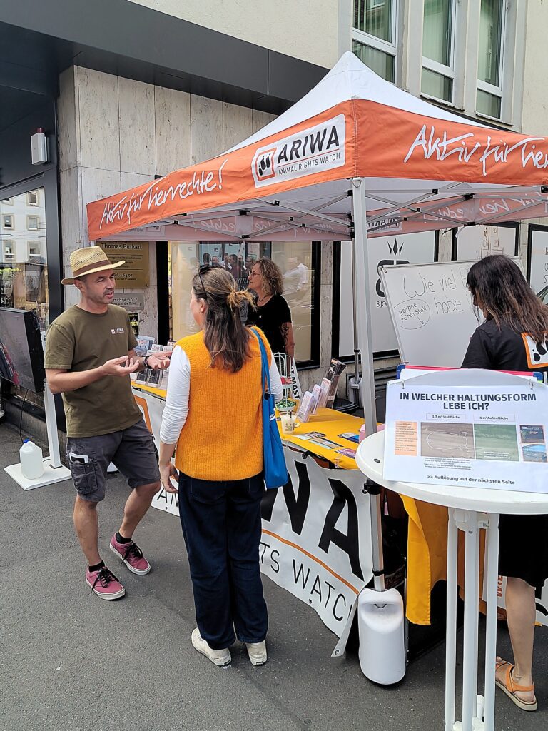 Ein Aktivist im ARIWA-T-Shirt und Strohhut spricht unter dem orangen Infostand-Pavillon mit einer Passantin in gelber Strickweste. Im Hintergrund weitere Aktivistinnen am Tisch, rechts ein Whiteboard mit der Frage „Wie viel Platz habe ich?" und die Haltungsform-Infotafel auf einem Stehtisch.