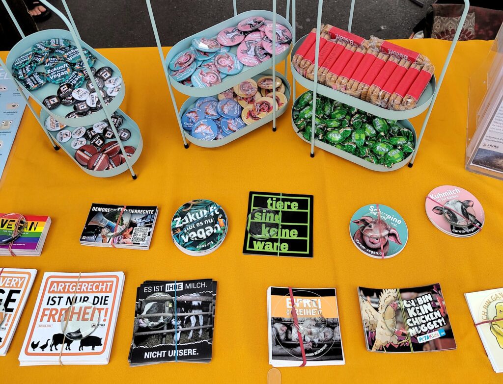 Draufsicht auf einen gelb gedeckten Tisch mit Werbeartikeln: mehrere Etagen mit Buttons zu Themen wie „Zukunft gibt es nur vegan", Kühen, Schweinen, Fischen und Hühnern, dazu Kekse und Bonbons. Darunter liegen Sticker und Flyer, u. a. „Artgerecht ist nur die Freiheit!", „Es ist ihre Milch. Nicht unsere." und „Ich bin kein Chicken Nugget!".