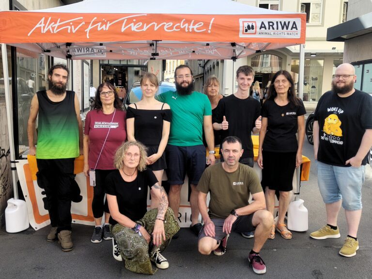 Zehn Aktivist:innen posieren gemeinsam für ein Gruppenfoto vor dem orangen ARIWA-Pavillon mit der Aufschrift „Aktiv für Tierrechte!". Mehrere tragen ARIWA-T-Shirts oder Shirts mit Slogans wie „Schlachthäuser schließen". Im Hintergrund eine Einkaufsstraße mit Geschäften.