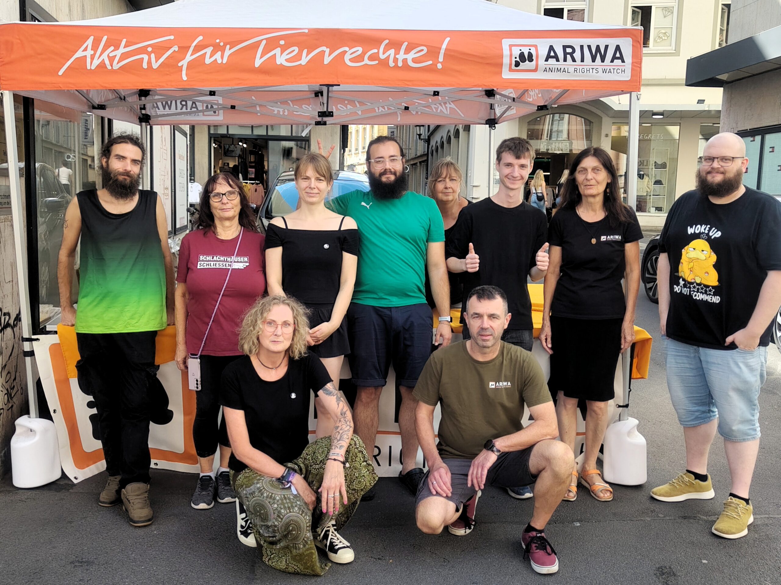 Zehn Aktivist:innen posieren gemeinsam für ein Gruppenfoto vor dem orangen ARIWA-Pavillon mit der Aufschrift „Aktiv für Tierrechte!". Mehrere tragen ARIWA-T-Shirts oder Shirts mit Slogans wie „Schlachthäuser schließen". Im Hintergrund eine Einkaufsstraße mit Geschäften.