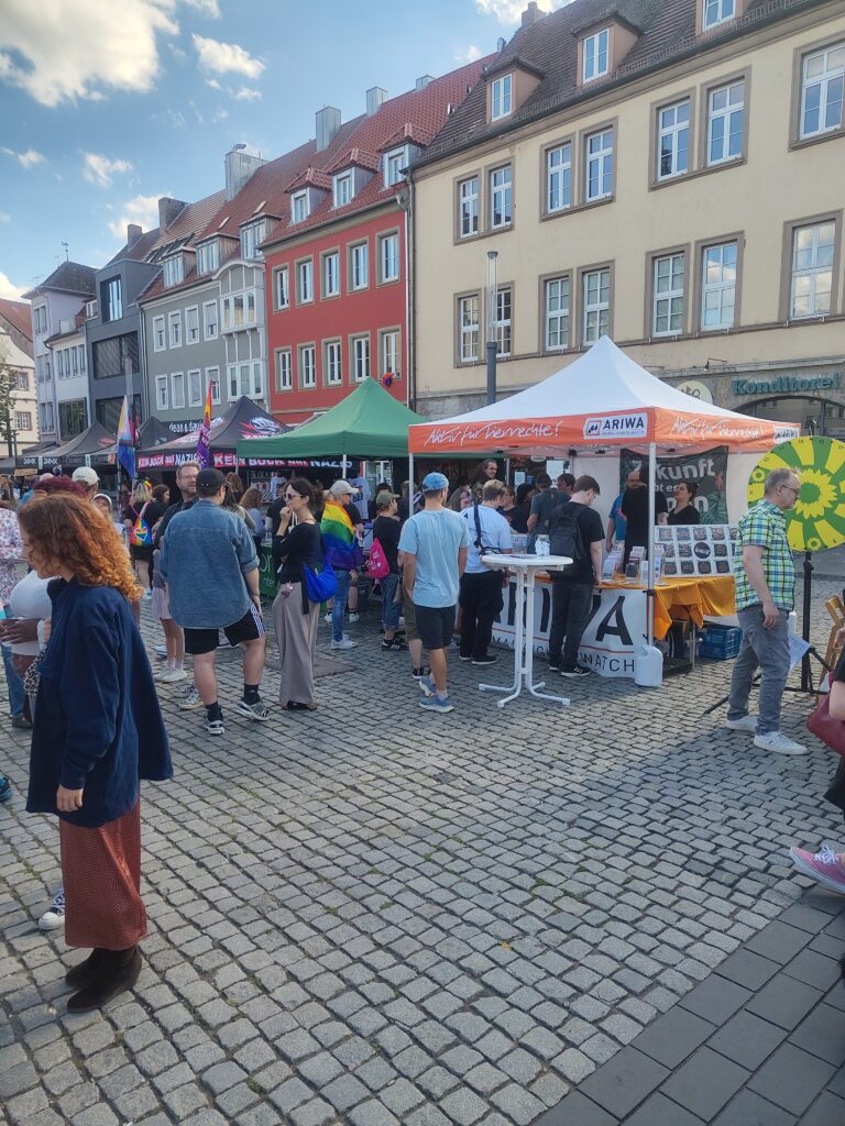 Weite Ansicht eines belebten Marktplatzes mit CSD-Ständen unter bunten Pavillons (grün, orange, schwarz) vor historischer Altstadtkulisse. Viele Besucher:innen stehen am ARIWA-Stand, eine Person trägt eine Regenbogenfahne um die Schultern. Rechts ein großes gelb-grünes Glücksrad.