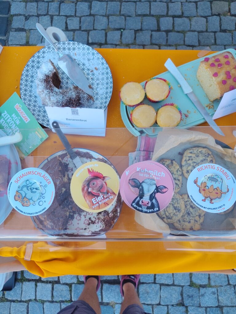 Draufsicht auf einen gelb gedeckten Kuchenbuffettisch: angeschnittener Bananenkuchen mit Puderzucker, Muffins, ein Zitronenkuchen mit pinker Glasur, Schokokuchen und Cookies. Zwischen den Kuchen liegen runde Sticker mit Cartoon-Motiven und Sprüchen wie „Eier sind zum Brüten da" (Huhn), „Kuhmilch ist für Kälber" (Kuh) und „Richtig stabil! Veganer Kuchen mit Apfelmus statt Ei".