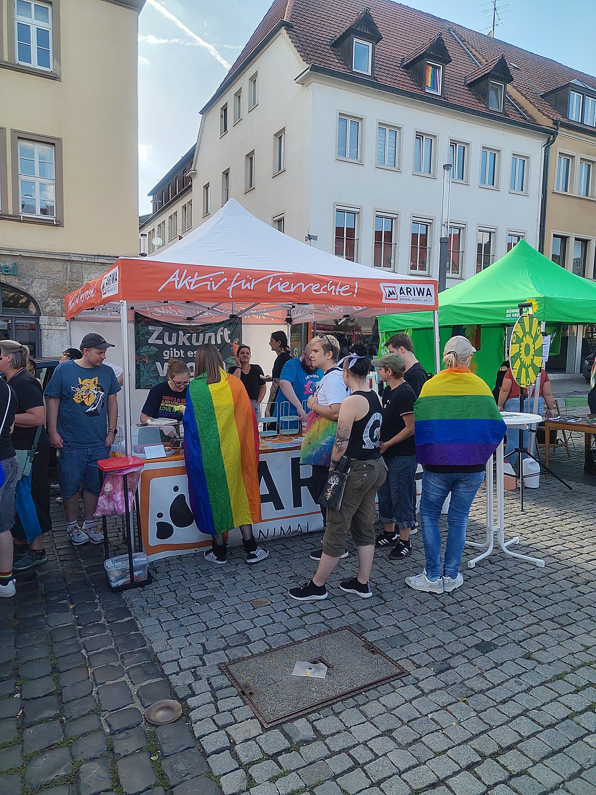 Gruppe von Menschen vor dem ARIWA-Stand, mehrere tragen Regenbogenfahnen als Umhang. Rechts das grüne Zelt von Bündnis 90/Die Grünen mit Glücksrad. Im Hintergrund ein Gebäude mit Regenbogenflagge im Dachfenster.