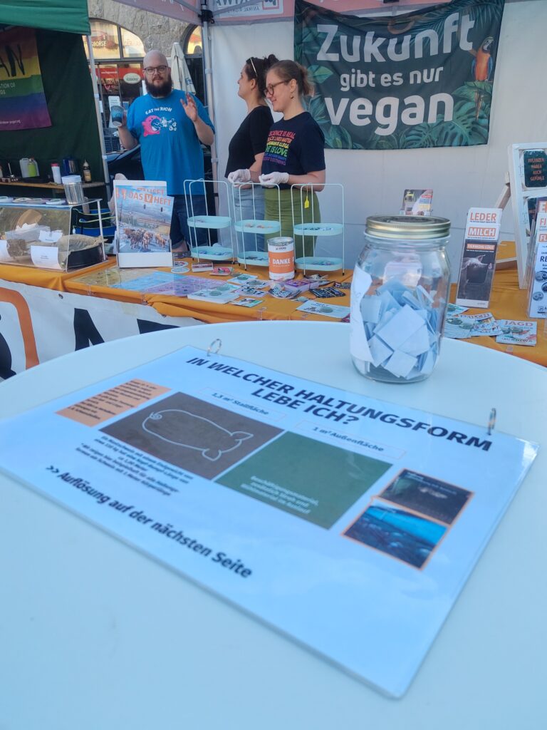 nnenansicht des ARIWA-Stands: Drei Aktivist:innen unterhalten sich hinter dem Verkaufstisch mit Buttons, Stickern und Broschüren wie „Das V-Heft". Im Hintergrund ein großes Banner „Zukunft gibt es nur vegan" mit Papageienmotiv. Im Vordergrund auf einem Stehtisch die Infotafel „In welcher Haltungsform lebe ich?"