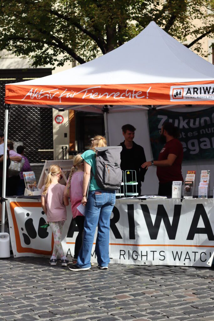ARIWA-Infostand mit orange-weißem Pavillon „Aktiv für Tierrechte!" unter einem Baum. Zwei Kinder und eine Frau mit Rucksack stehen am Tisch, dahinter zwei Aktivisten im Gespräch. Auf dem Tisch liegen Broschüren, im Hintergrund ein Banner „Zukunft gibt es nur vegan".