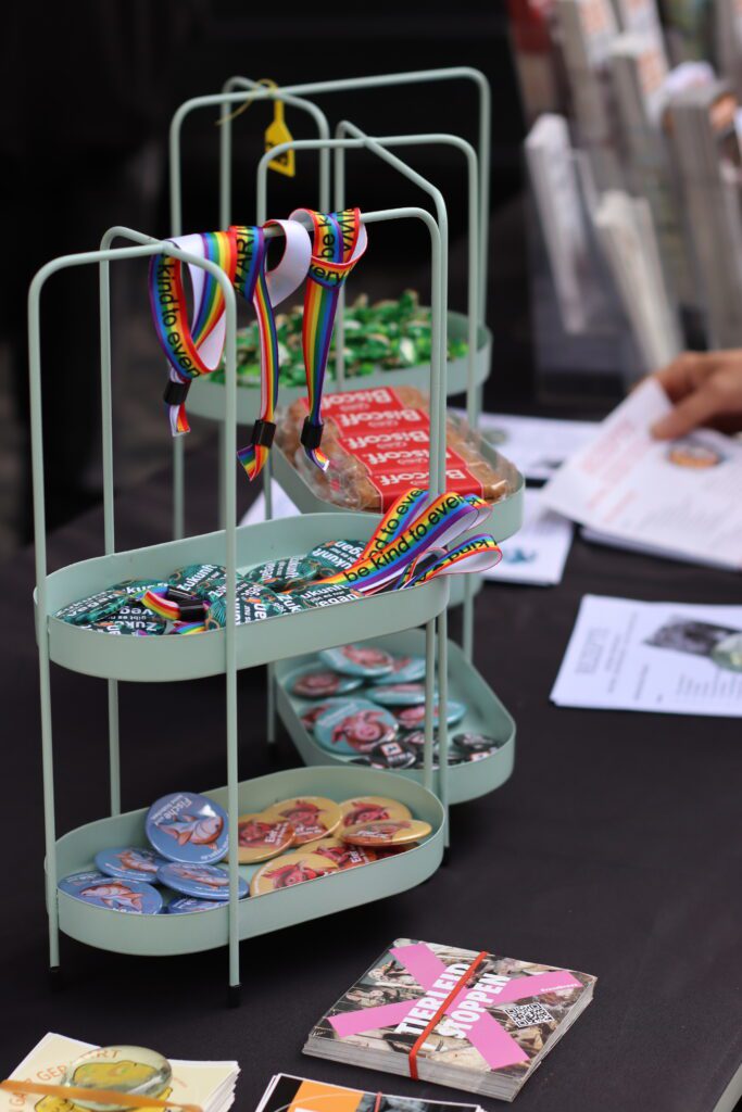 Nahaufnahme eines Etagenständers mit ARIWA-Merchandise: Buttons mit Motiven von Fischen, Hühnern und Schweinen, dazu Kekse und Regenbogenbändchen mit der Aufschrift „be kind to every kind". Im Hintergrund liegen Flyer, darunter einer mit „Tierleid stoppen".