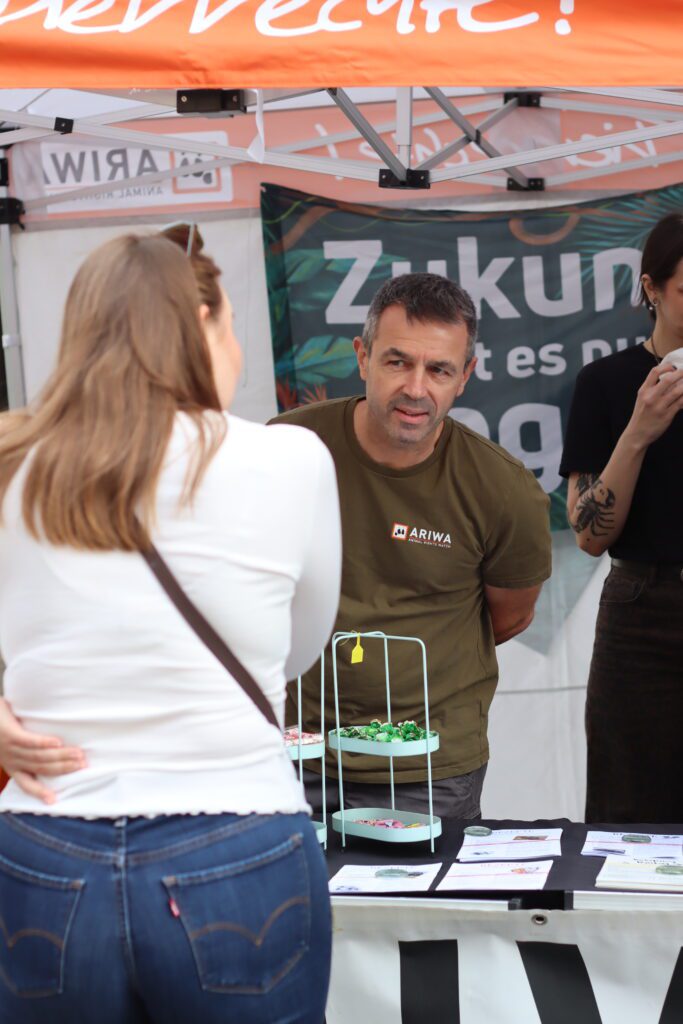 Ein Aktivist im olivgrünen ARIWA-T-Shirt spricht unter dem Pavillon mit einer Besucherin, im Hintergrund das Banner „Zukunft gibt es nur vegan" und eine weitere Aktivistin mit tätowiertem Arm. Auf dem Tisch ein Etagenständer mit Süßigkeiten.