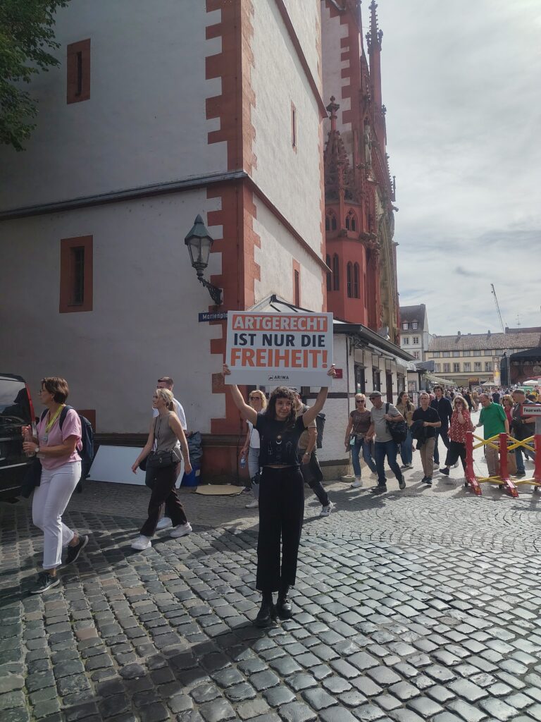Eine Aktivistin steht allein vor der Marienkapelle am Marienplatz und hält ein Schild hoch: „Artgerecht ist nur die Freiheit!" mit ARIWA-Logo. Im Hintergrund strömen viele Festbesucher:innen vorbei, rechts eine Absperrung mit „Feuerwehr"-Schild.